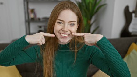 Photo for A cheerful young caucasian woman smiles, pointing to her mouth, in a cozy living room setting. - Royalty Free Image