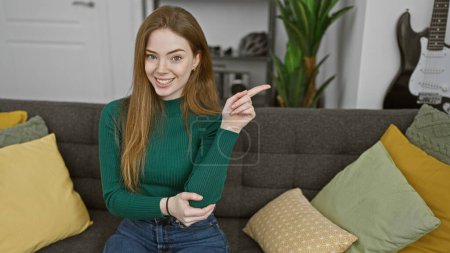 Photo for Smiling young blonde woman pointing while seated on a sofa in a cozy living room with colorful pillows. - Royalty Free Image