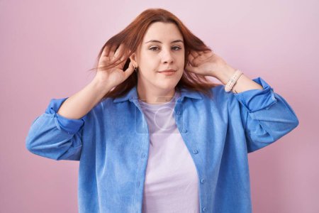 Photo for Young hispanic woman with red hair standing over pink background trying to hear both hands on ear gesture, curious for gossip. hearing problem, deaf - Royalty Free Image