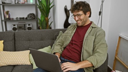Smiling young hispanic man with glasses and a beard comfortably using a laptop on a couch in a cozy apartment living room.