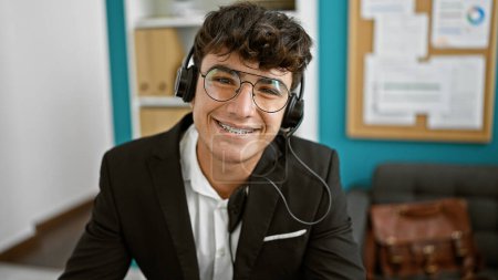 Photo for Confident young hispanic teenager working in business, wearing headset handling customer service with a smile inside his office - Royalty Free Image