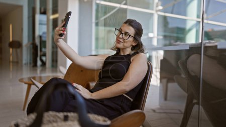 Foto de Mujer hispana segura con gafas divirtiéndose, tomando una selfie radiante mientras se sienta casualmente en un moderno salón de hotel - Imagen libre de derechos
