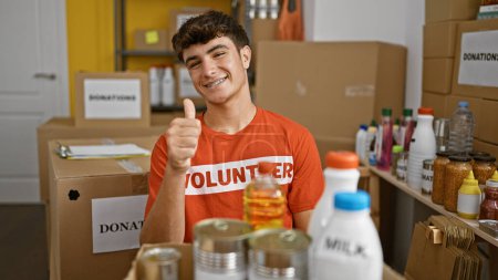 Photo for Confident young hispanic teenager smiling, giving a thumbs up in acknowledgment of great work while volunteering at a lively charity center - Royalty Free Image