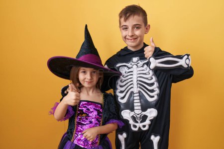 Cheerful brother-sister duo wearing halloween costumes, giving cool 'ok' signs, beaming with joy and confidence on isolated yellow background