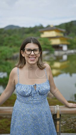 Téléchargez les photos : Attrayant hispanique femme, joie rayonnant de son sourire rayonnant, se tient confiant et décontracté dans ses lunettes à couper le souffle kinkaku-ji pavillon d'or, un bijou de kyoto, Japon - en image libre de droit