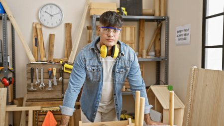 Photo for Serious-faced young hispanic man, a professional carpenter, wearing headphones and glasses craftily working in his indoor carpentry workshop - Royalty Free Image