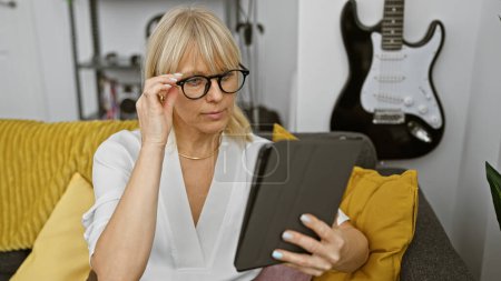 Foto de Una mujer rubia enfocada con gafas leyendo una tableta en una sala de estar moderna con un fondo de guitarra - Imagen libre de derechos