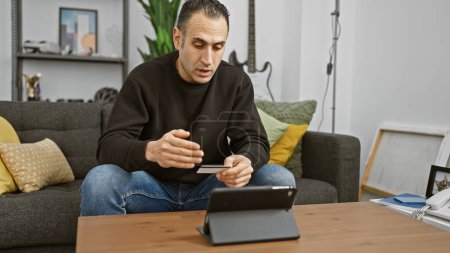 Photo for A focused hispanic man uses a credit card while browsing on a tablet in a cozy living room setting. - Royalty Free Image