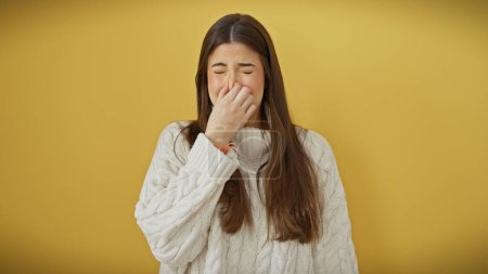 Foto de Mujer hispana joven pellizcándose la nariz para evitar el mal olor, aislada sobre fondo amarillo. - Imagen libre de derechos