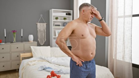 Foto de Hombre de mediana edad en pijama aparece angustiado en un dormitorio moderno, de pie junto a la cama con pesas de ejercicio. - Imagen libre de derechos