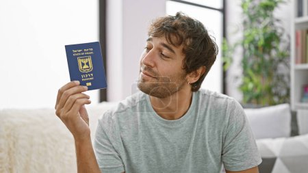 Photo for Confident, bearded young man joyfully flashing his israeli passport, casually sitting on the sofa at his cozy apartment living room, ready for a fun-filled holiday trip! - Royalty Free Image