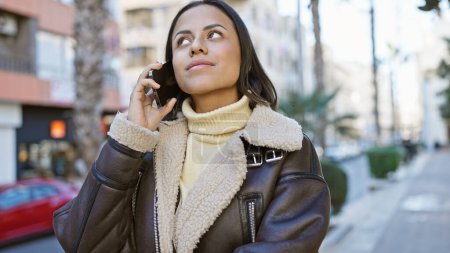 Eine stilvolle hispanische Frau telefoniert, strahlt Zuversicht aus, während sie in einer städtischen Straße spaziert.