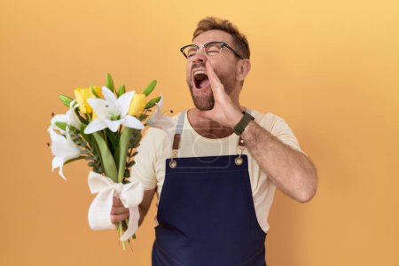 Foto de Hombre de mediana edad con floristería barba sosteniendo flores gritando y gritando fuerte a un lado con la mano en la boca. concepto de comunicación. - Imagen libre de derechos