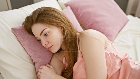 Photo for A young caucasian woman, with blonde hair, lies pensively in bed within the intimate setting of a softly-lit bedroom. - Royalty Free Image