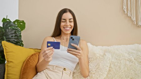 Photo for Confident, smiling young hispanic woman joyfully shopping from her cozy living room sofa, making payments online with her credit card and smartphone. - Royalty Free Image