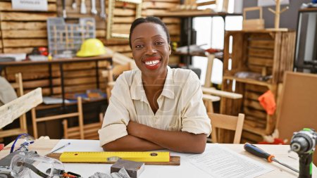 Foto de Sonriente mujer afroamericana carpintera, confiada y relajada, increíblemente atractiva con los brazos cruzados, sentada en la mesa de carpintería en el taller interior - Imagen libre de derechos