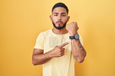 Téléchargez les photos : Jeune homme hispanique debout sur fond jaune pressé pointant vers regarder le temps, l'impatience, regardant la caméra avec une expression détendue - en image libre de droit