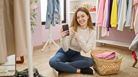 Foto de Una joven mujer caucásica sonriente apuntando a un teléfono inteligente en una habitación de ropa con una cesta de ropa - Imagen libre de derechos