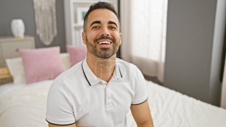 Téléchargez les photos : Jeune homme hispanique souriant assis avec désinvolture sur un lit dans un intérieur de chambre bien décoré, évoquant un sentiment de confort et de détente. - en image libre de droit
