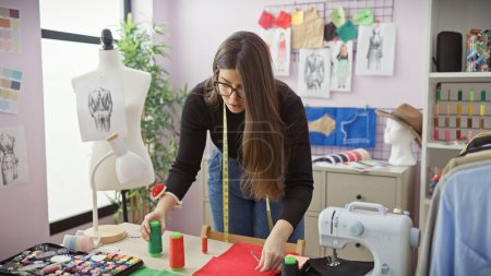Foto de Una mujer enfocada en tela de medida de sastre en un colorido atelier rodeado de equipos de costura y maniquíes. - Imagen libre de derechos