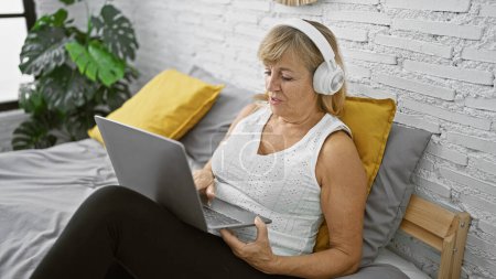 Photo for Relaxed middle age blonde woman enjoying morning tunes, beautifully sitting in her bedroom, laptop on her lap, headphones on, her serious face glowing in the soft indoor light. - Royalty Free Image