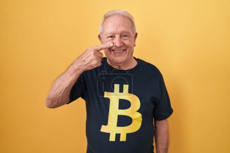 Foto de Hombre mayor con el pelo gris con camiseta bitcoin apuntando con el dedo de la mano a la cara y la nariz, sonriendo alegre. concepto de belleza - Imagen libre de derechos
