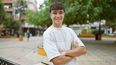 Photo for Cheerful young hispanic teenager, beaming with confidence, expressing joy through a playful arm crossed gesture in the heart of a lush green city park - Royalty Free Image