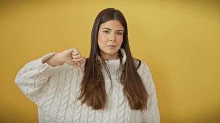 Foto de Una joven hispana hace un gesto de pulgares hacia abajo contra un fondo amarillo aislado, sugiriendo insatisfacción. - Imagen libre de derechos