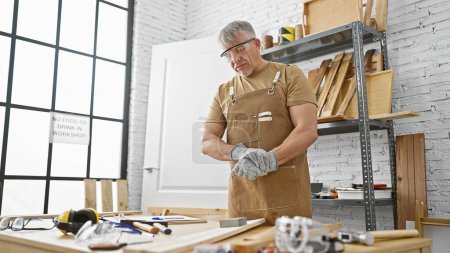 Foto de Un hombre maduro con gafas y un delantal se levanta pensativamente en un taller de carpintería rodeado de madera y herramientas. - Imagen libre de derechos