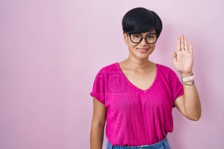 Foto de Joven mujer asiática con pelo corto de pie sobre fondo rosa renunciando a decir hola feliz y sonriente, gesto de bienvenida amistoso - Imagen libre de derechos