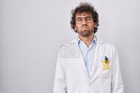 Photo for Hispanic young man wearing doctor uniform puffing cheeks with funny face. mouth inflated with air, crazy expression. - Royalty Free Image