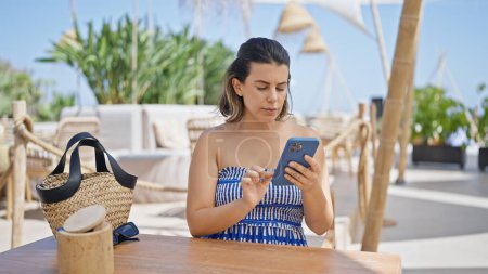 Téléchargez les photos : Jeune femme hispanique utilisant smartphone assis sur la table à la terrasse ensoleillée du restaurant - en image libre de droit