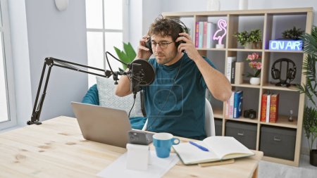 Téléchargez les photos : Bel homme hispanique réglage casque dans un studio de radio moderne avec ordinateur portable et microphone. - en image libre de droit