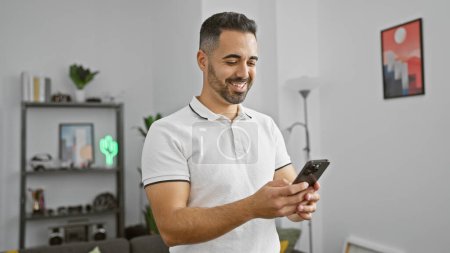 Photo for Smiling young hispanic man holding smartphone in modern living room - Royalty Free Image