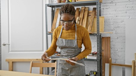 Foto de Mujer afroamericana con trenzas con gafas de seguridad lee planes en taller de carpintería - Imagen libre de derechos