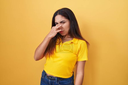 Téléchargez les photos : Jeune femme arabe debout sur fond jaune sentant quelque chose de puant et dégoûtant, odeur intolérable, retenant la respiration avec les doigts sur le nez. mauvaise odeur - en image libre de droit