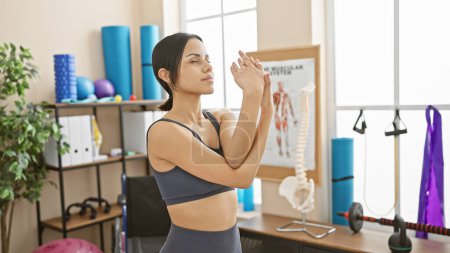 Photo for A young hispanic woman in workout attire stretches her arms in a well-equipped physiotherapy clinic room. - Royalty Free Image