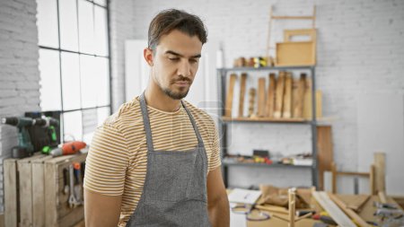 Photo for Handsome hispanic man with a beard wearing an apron in a well-lit carpentry workshop - Royalty Free Image