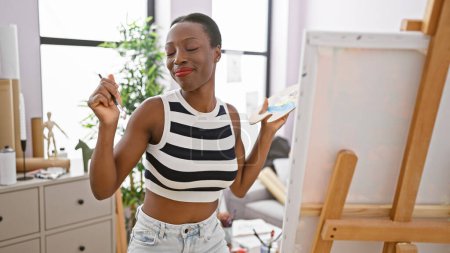 Téléchargez les photos : Artiste afro-américaine confiante, dansant et souriant dans un studio d'art tout en appréciant et apprenant la peinture lors d'un cours de créativité - en image libre de droit