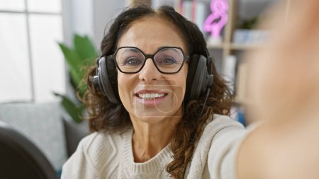 Téléchargez les photos : Femme hispanique dans la cinquantaine, prenant un selfie à l'intérieur avec écouteurs, souriant à la caméra. - en image libre de droit
