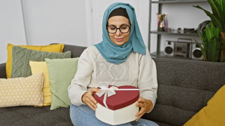 Photo for A mature woman wearing a hijab smiles while holding a heart-shaped gift box in a cozy living room. - Royalty Free Image