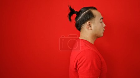 Téléchargez les photos : Cool, détendu jeune homme chinois sérieusement regarder sur le côté contre le mur rouge isolé, exprimant mode de vie concentré avec coiffure asiatique décontractée - en image libre de droit