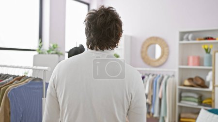 Photo for Rear view of a young man in a white shirt standing in a tidy, well-lit dressing room filled with clothes - Royalty Free Image