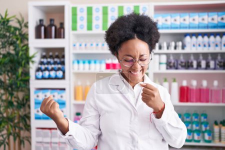 Téléchargez les photos : Belle femme africaine aux cheveux bouclés travaillant à la pharmacie pharmacie très heureuse et excitée de faire geste gagnant avec les bras levés, souriant et criant pour le succès. concept de célébration. - en image libre de droit