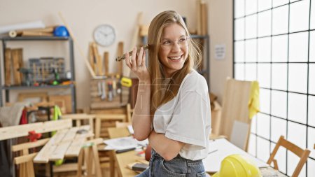Téléchargez les photos : Souriante jeune femme tenant un crayon dans un atelier de menuiserie ensoleillé - en image libre de droit