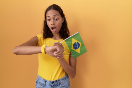 Téléchargez les photos : Jeune femme hispanique tenant drapeau brésilien regardant le temps de la montre inquiet, peur de se faire tard - en image libre de droit
