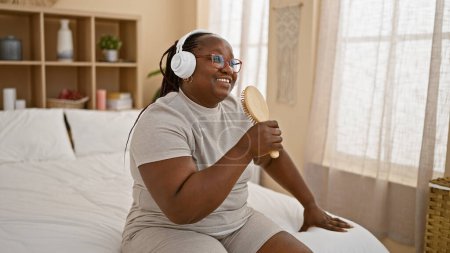 Téléchargez les photos : Femme afro-américaine confiante profitant du matin, se détendre sur le lit en chantant une chanson à l'aide d'une brosse comme un microphone, entendre sa voix à travers des écouteurs dans sa chambre confortable - en image libre de droit