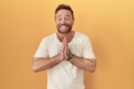 Middle age man with beard standing over yellow background praying with hands together asking for forgiveness smiling confident. 