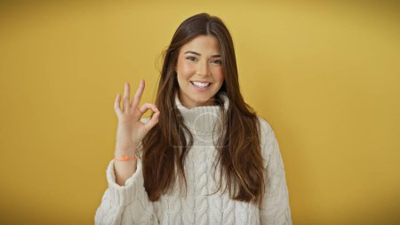 Foto de Una joven hispana sonriente dando una señal de ok en un suéter blanco, aislada sobre un fondo amarillo. - Imagen libre de derechos