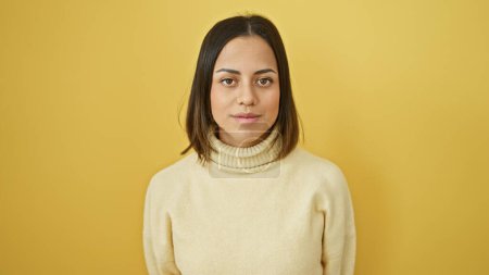 Foto de Retrato de una joven hispana con expresión neutra, de pie sobre un fondo amarillo aislado. - Imagen libre de derechos